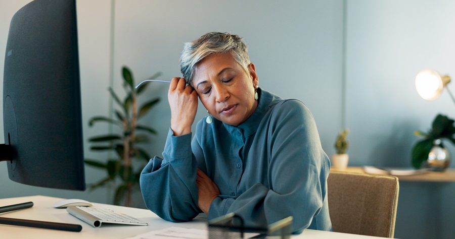 Older adult woman sitting at her desk with her eyes close and her hand on the side of her head.