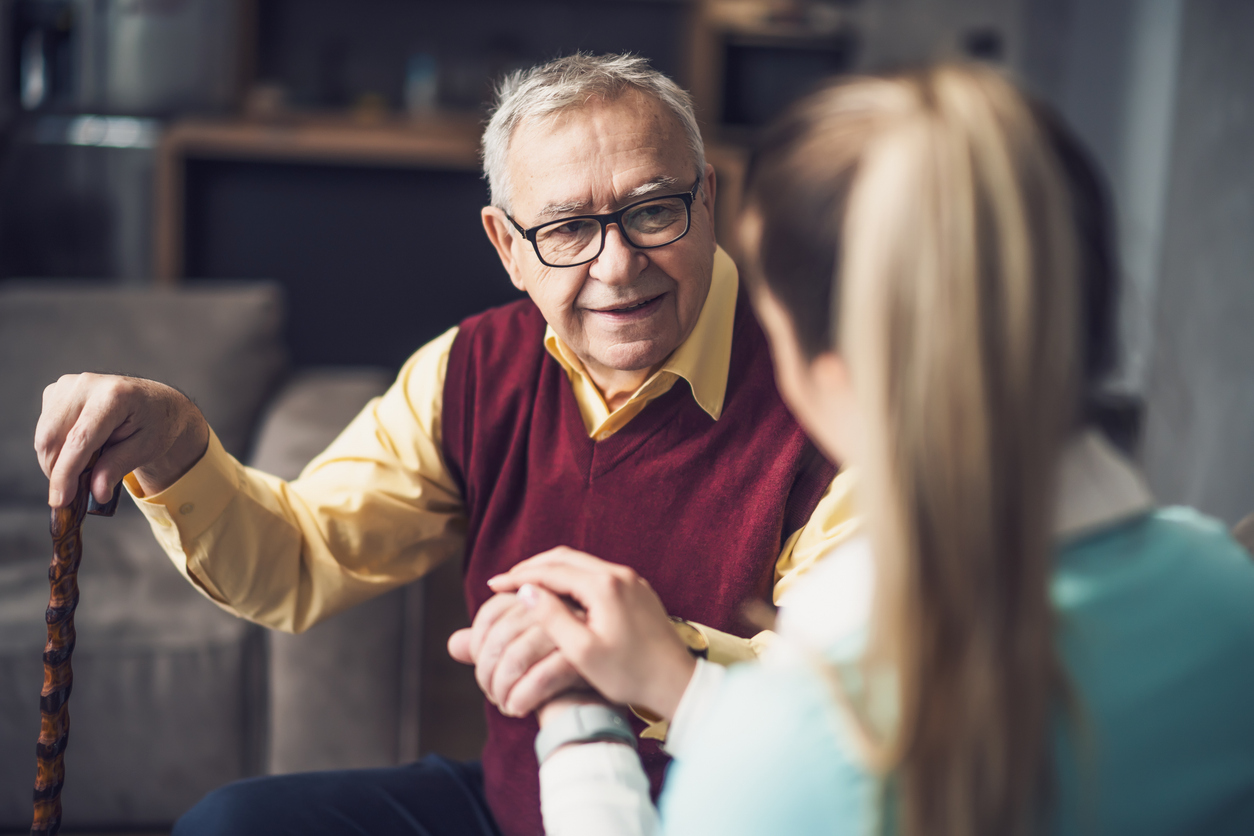 Caregiver holding the hand of an elderly man sitting on a couch with a cane.