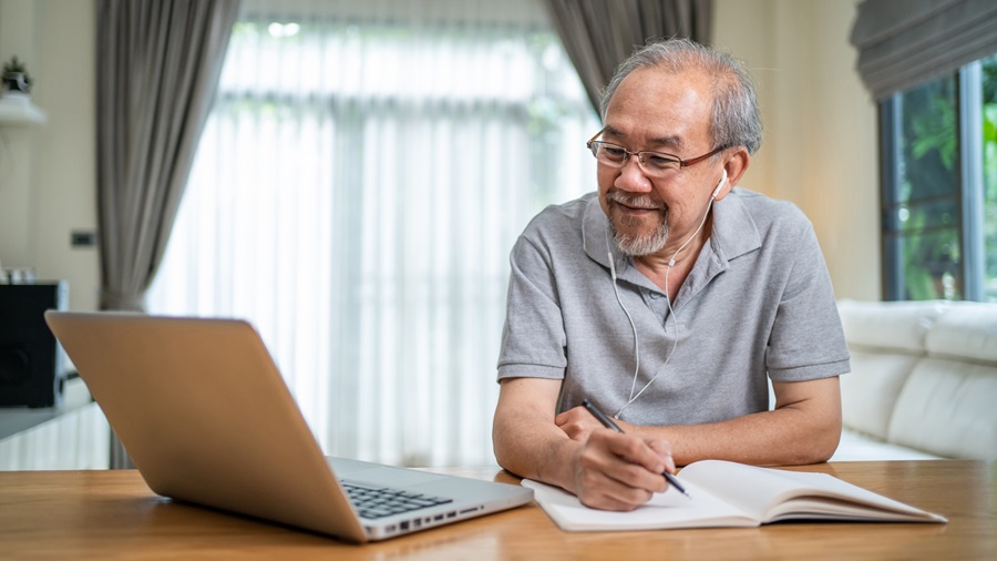 Older adult with headphones on and writing in a journal while watching something on a laptop.