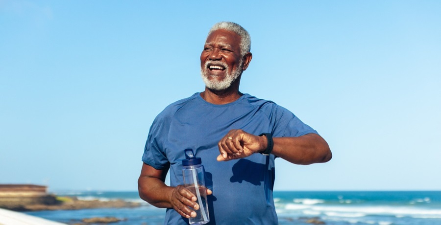 Elderly male holding a water bottle and checking his smartwatch while walking near a beach.