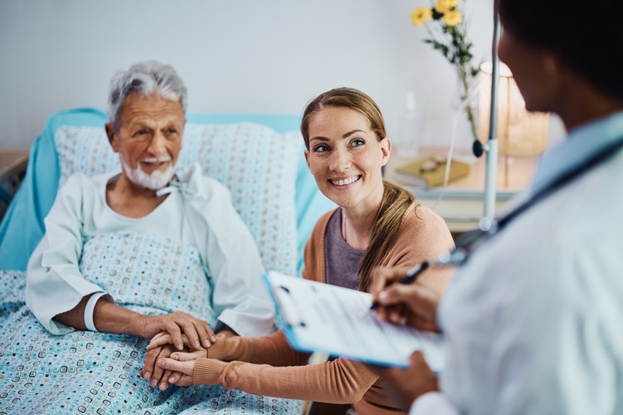 Older adult sitting in a hospital bed while holding hands with their caregiver and speaking to their doctor.