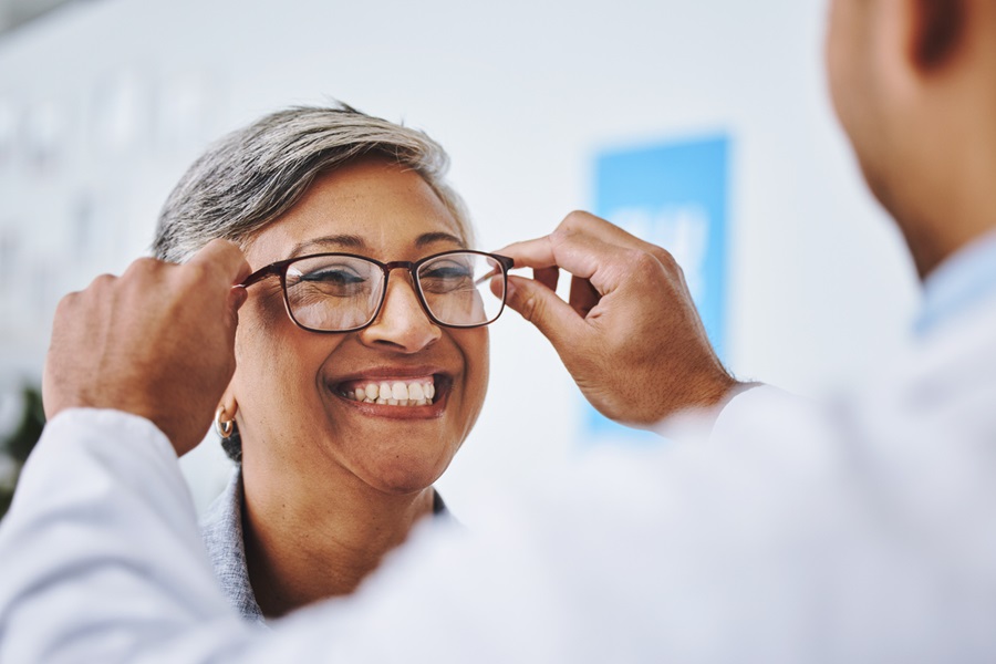 Woman smiling as an eye doctor places eyeglasses on her face.
